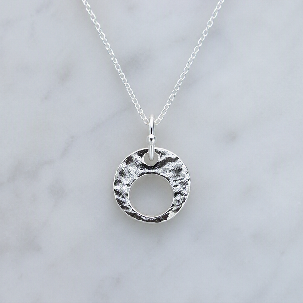 A silver pendant necklace with a textured, hammered circular disc design on a delicate link chain, displayed on a light gray marble surface. A small 'Made with AI' label is in the top right corner.
